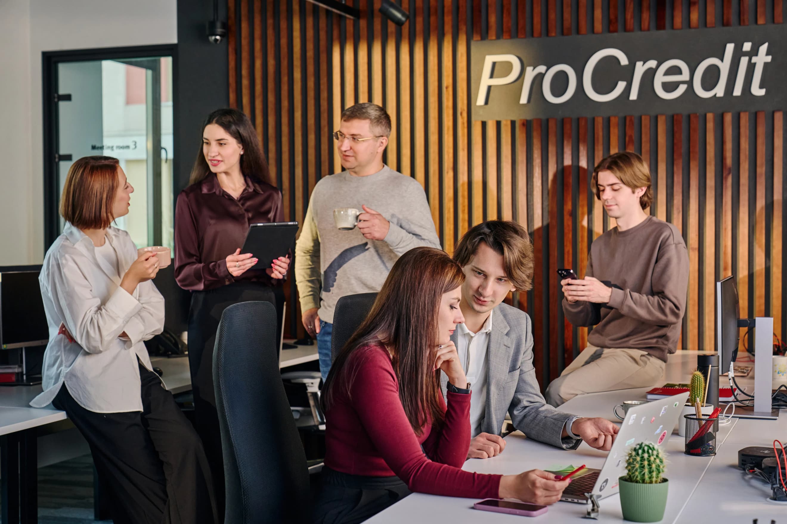 Шестеро людей спілкуються в офісному приміщенні. Двоє сидять і працюють за ноутбуком, решта стоять поряд і спілкуються