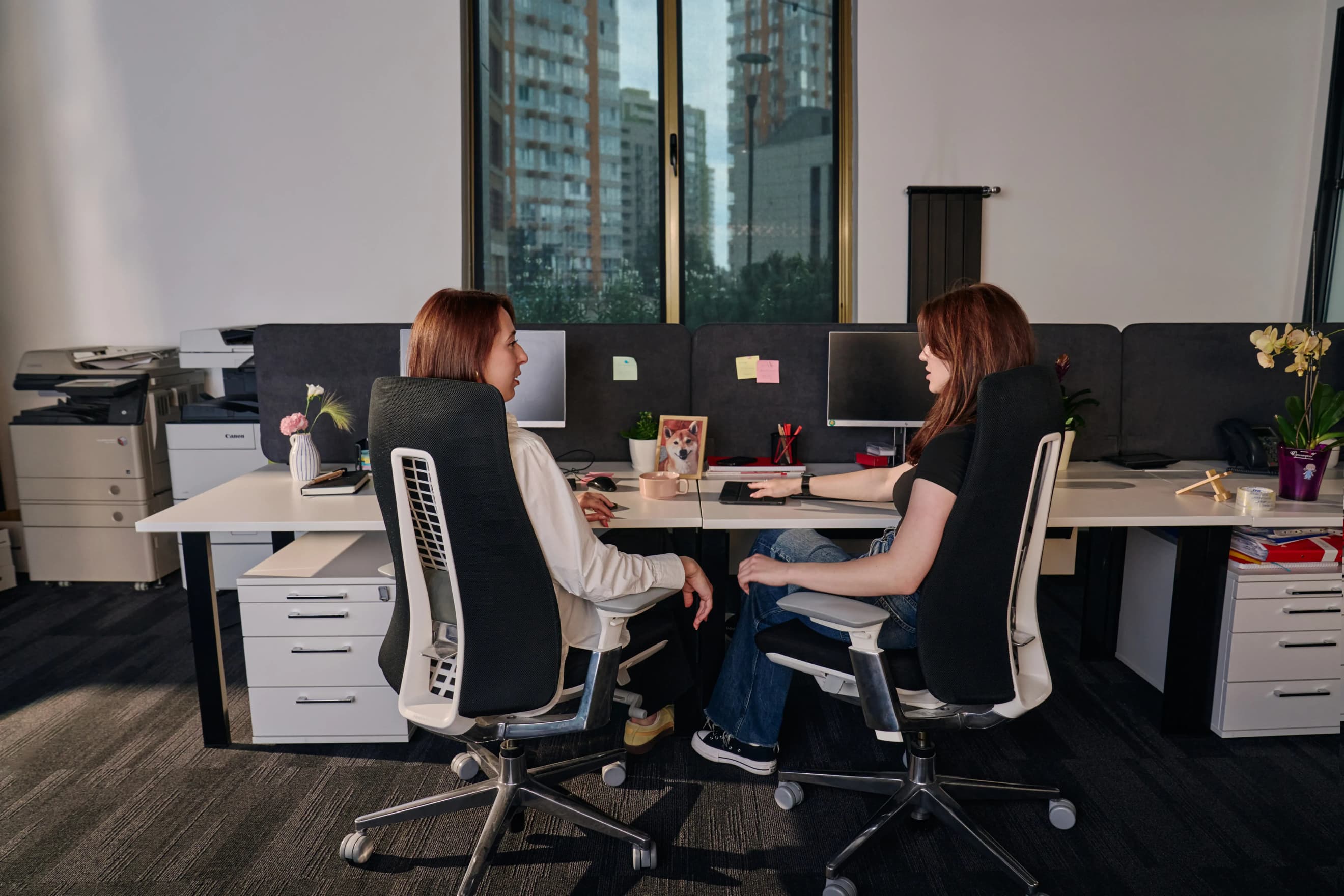 Дві жінки сидять в офісних кріслах за столом з ноутбуками біля вікна з краєвидом міста