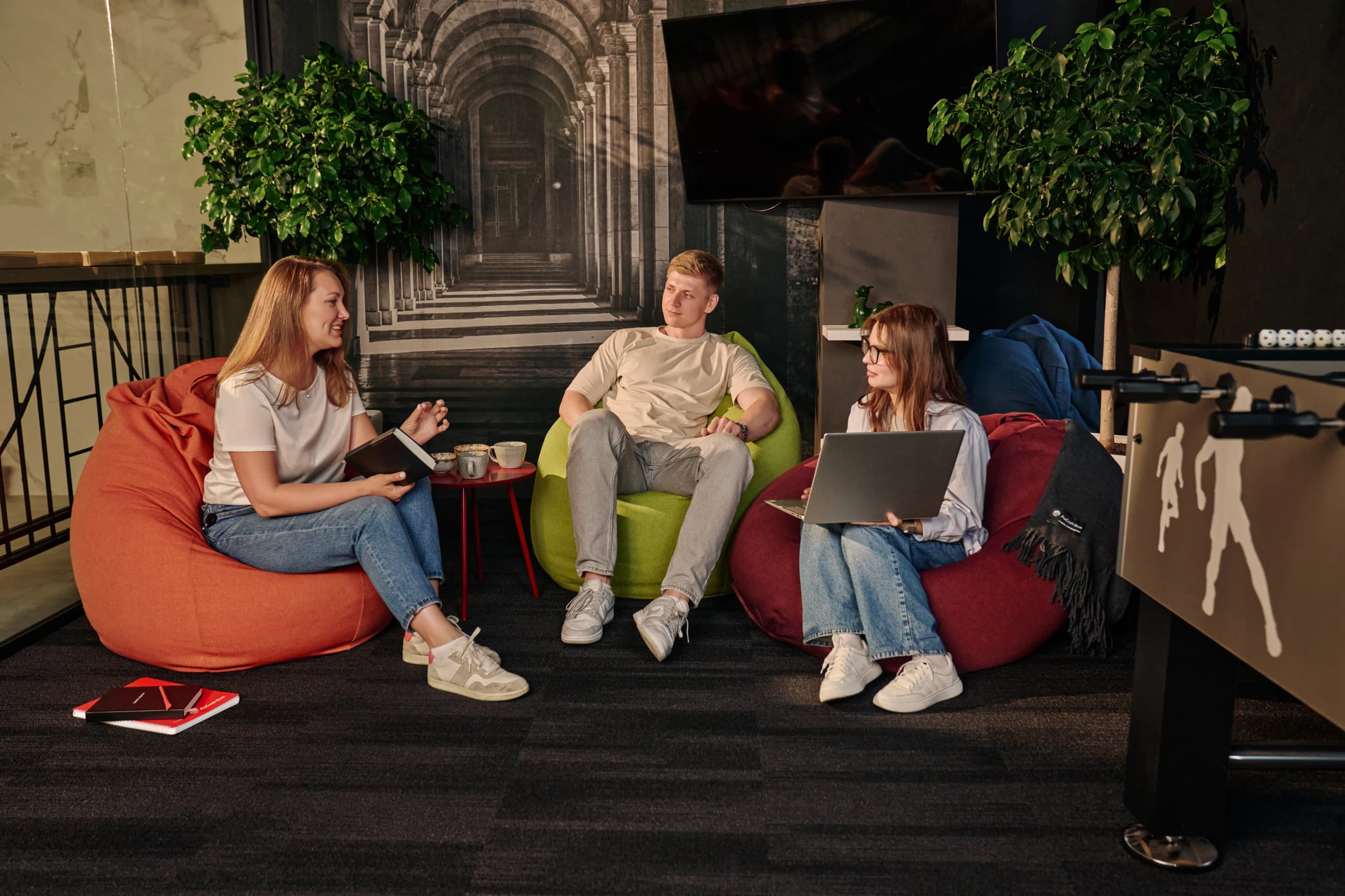Дві жінки і чоловік сидять на м’яких кріслах у зоні відпочинку з ноутбуком, блокнотом і столиком з напоями