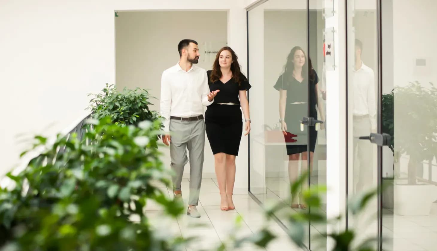 Чоловік і жінка йдуть коридором офісу повз скляні двері та спілкуються 