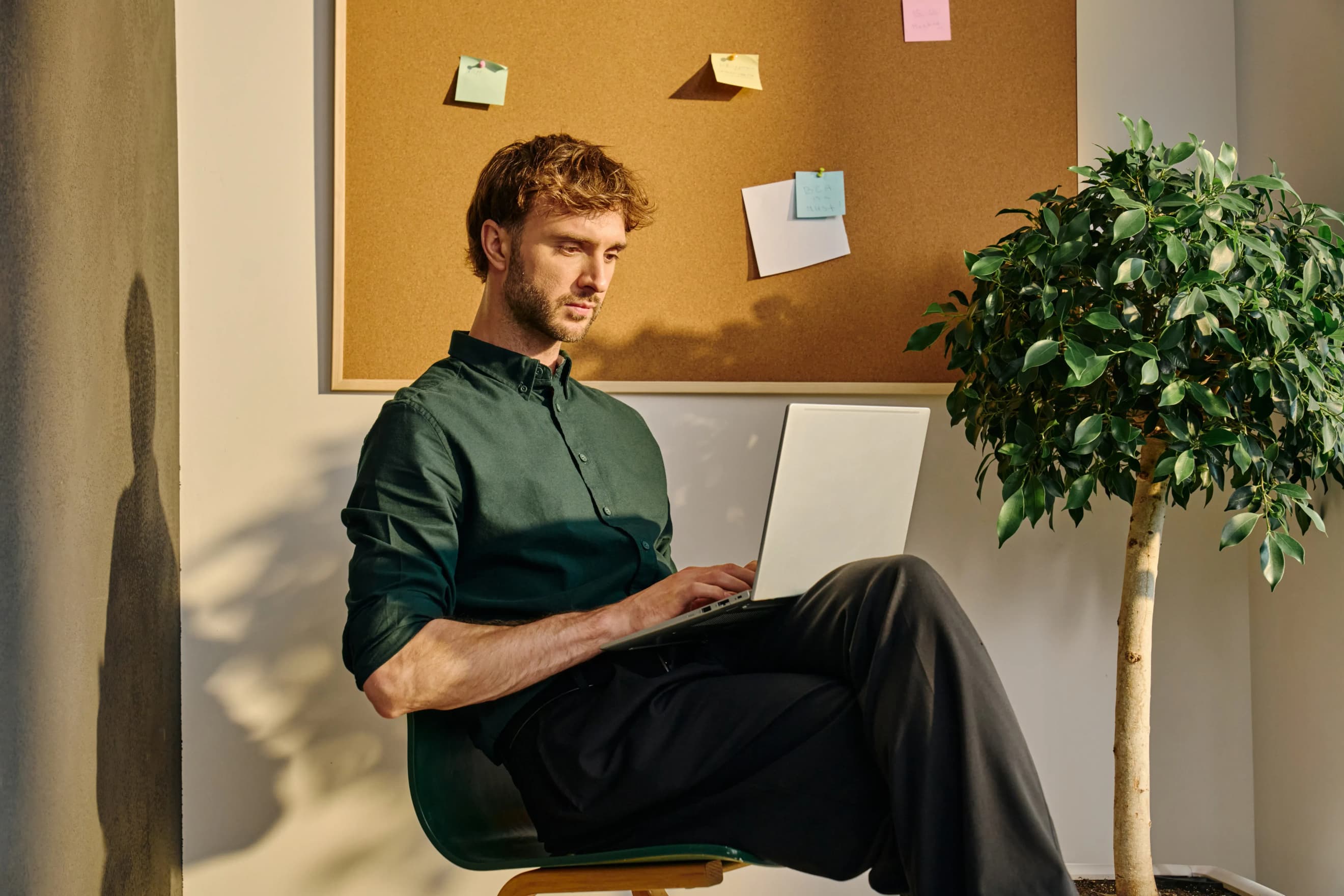 Чоловік сидить на стільці з ноутбуком на колінах біля дошки з наліпками та поруч із кімнатною рослиною