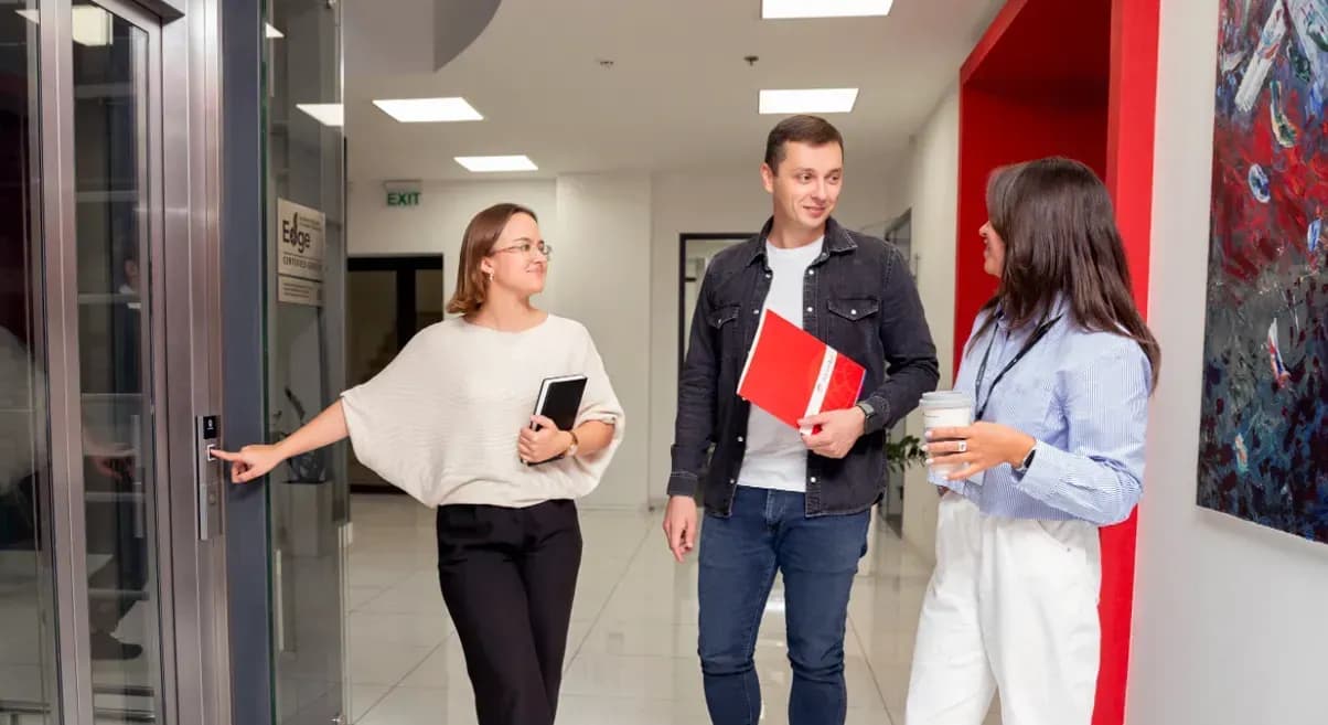 Дві жінки і чоловік стоять у коридорі офісу біля ліфту з блокнотом, папкою з документами та чашкою з напоєм