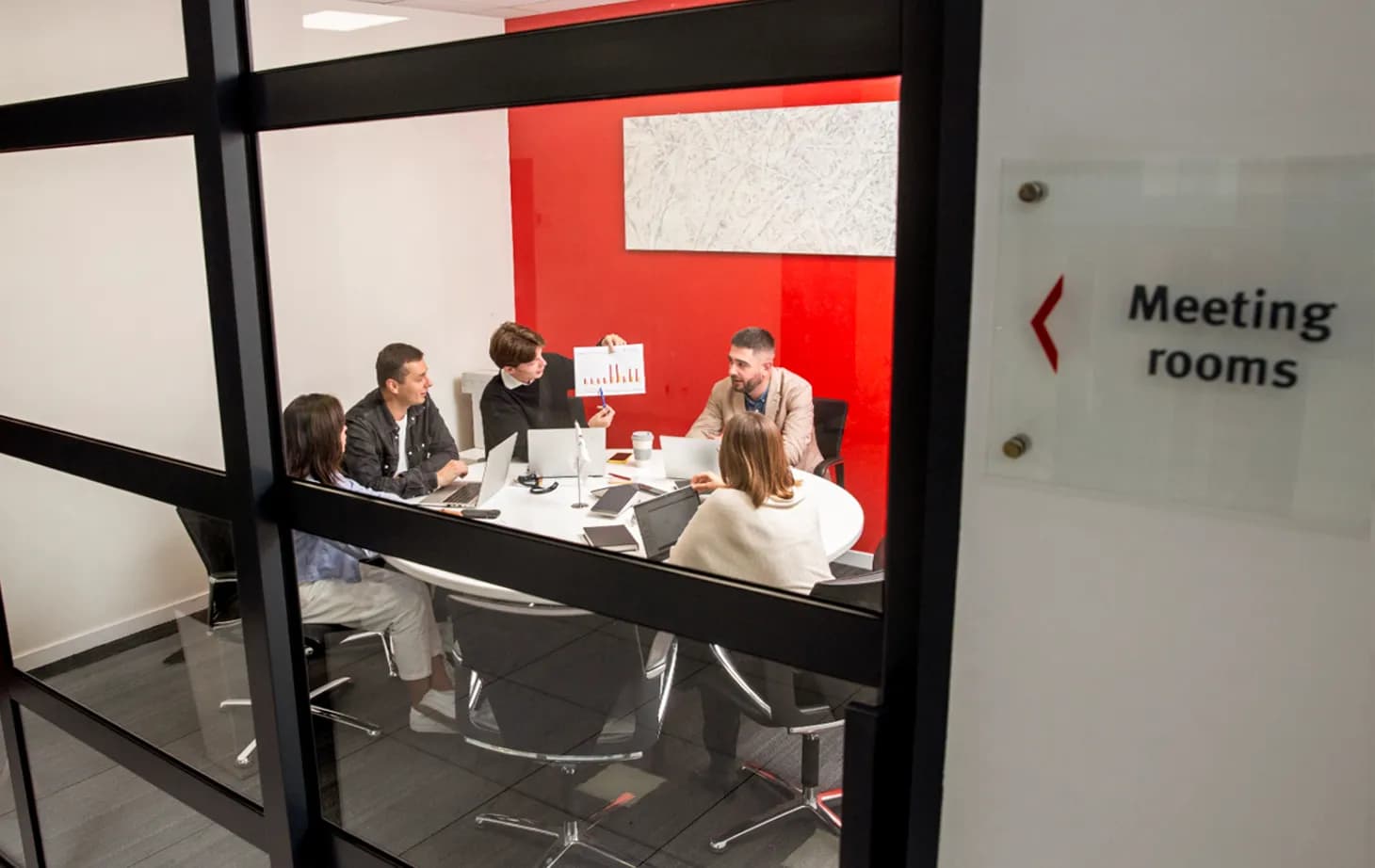 Крізь скляні двері кімнати видно групу людей, яка сидить з ноутбуками за круглим столом і обговорює роздрукований графік