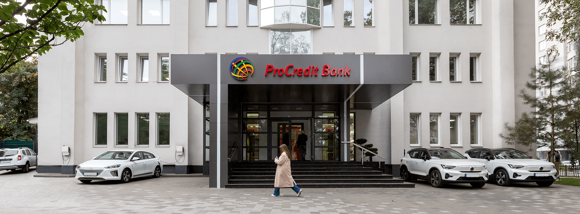 Вхід до ПроКредит Банку із темно-сірим навісом, сходами та скляними дверима, поруч припарковані електроавтомобілі