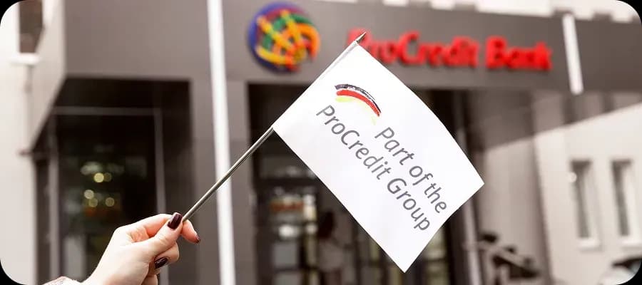 Рука тримає прапорець із написом «Part of the ProCredit Group» на тлі будівлі ProCredit Bank