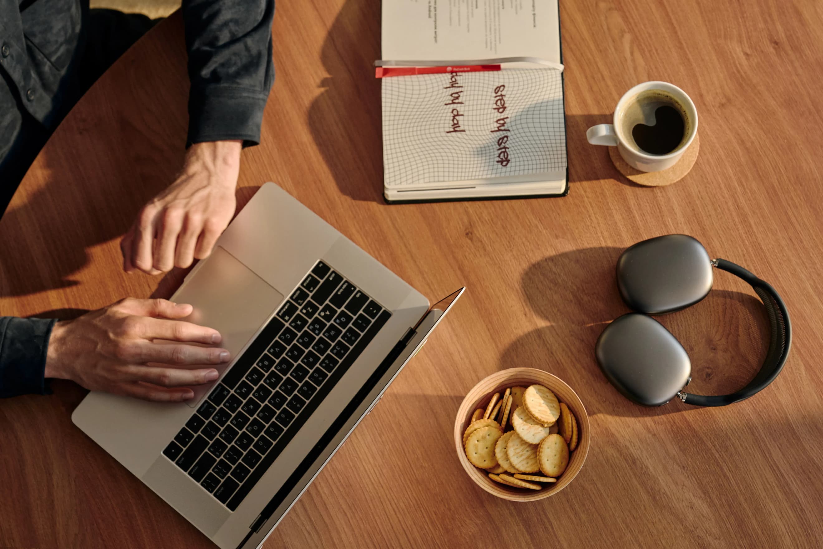 Робочий стіл із ноутбуком, блокнотом, навушниками, горнятком напою і тарілкою з крекерами