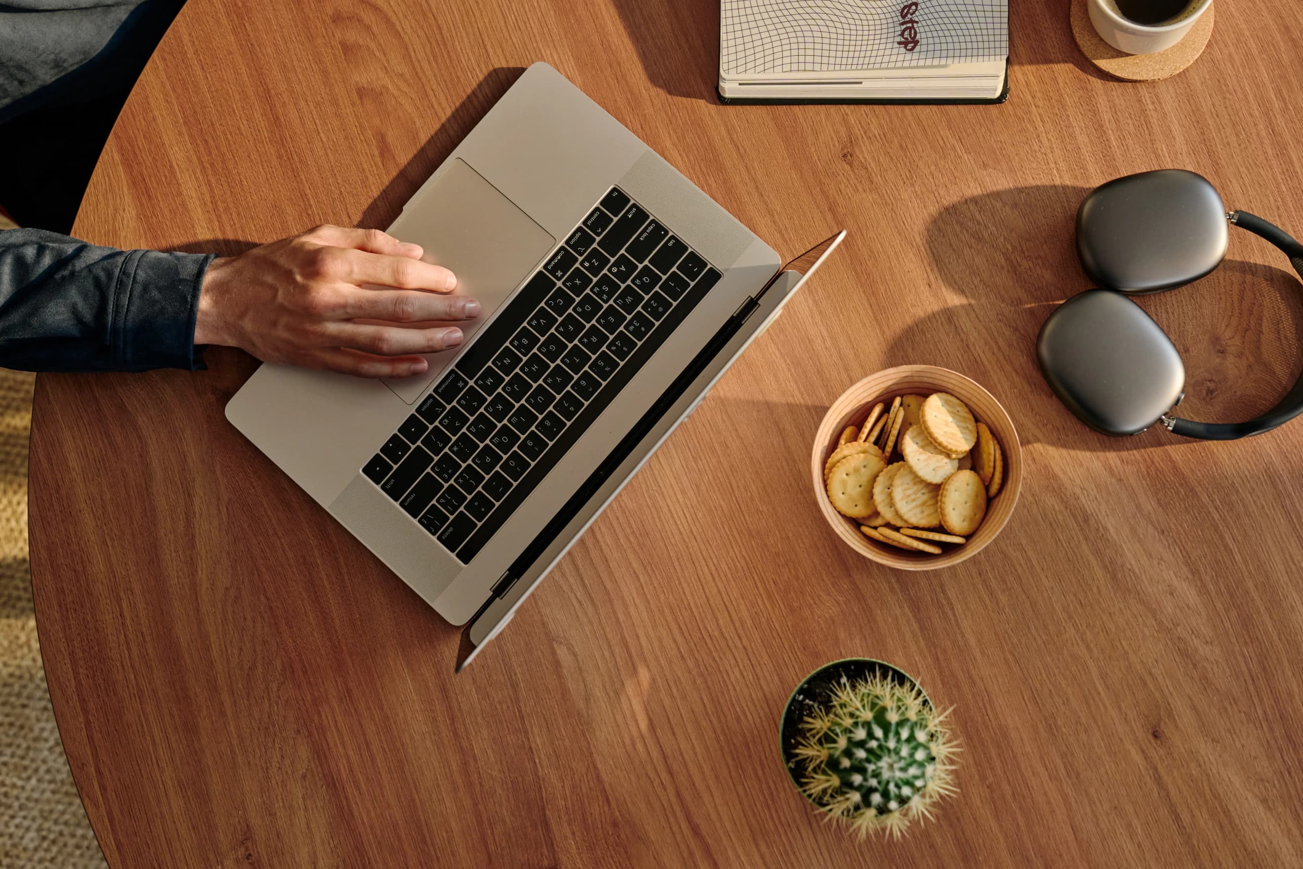 Рука працює за ноутбуком на дерев’яному столі поруч із навушниками, блокнотом, кактусом і тарілкою з крекерами.