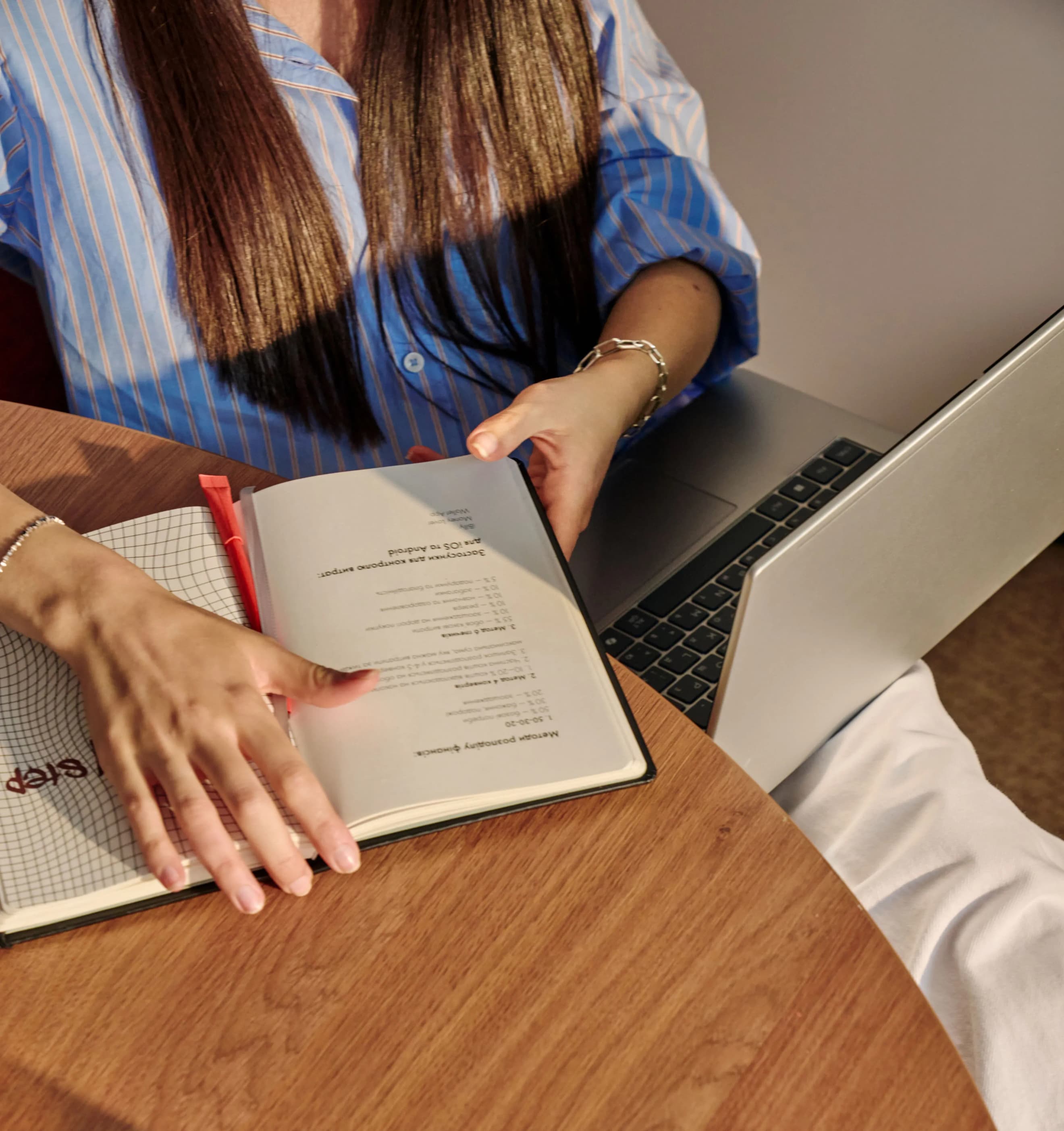 Жінка переглядає записи у відкритому блокноті, тримаючи ручку біля ноутбука на дерев’яному столі