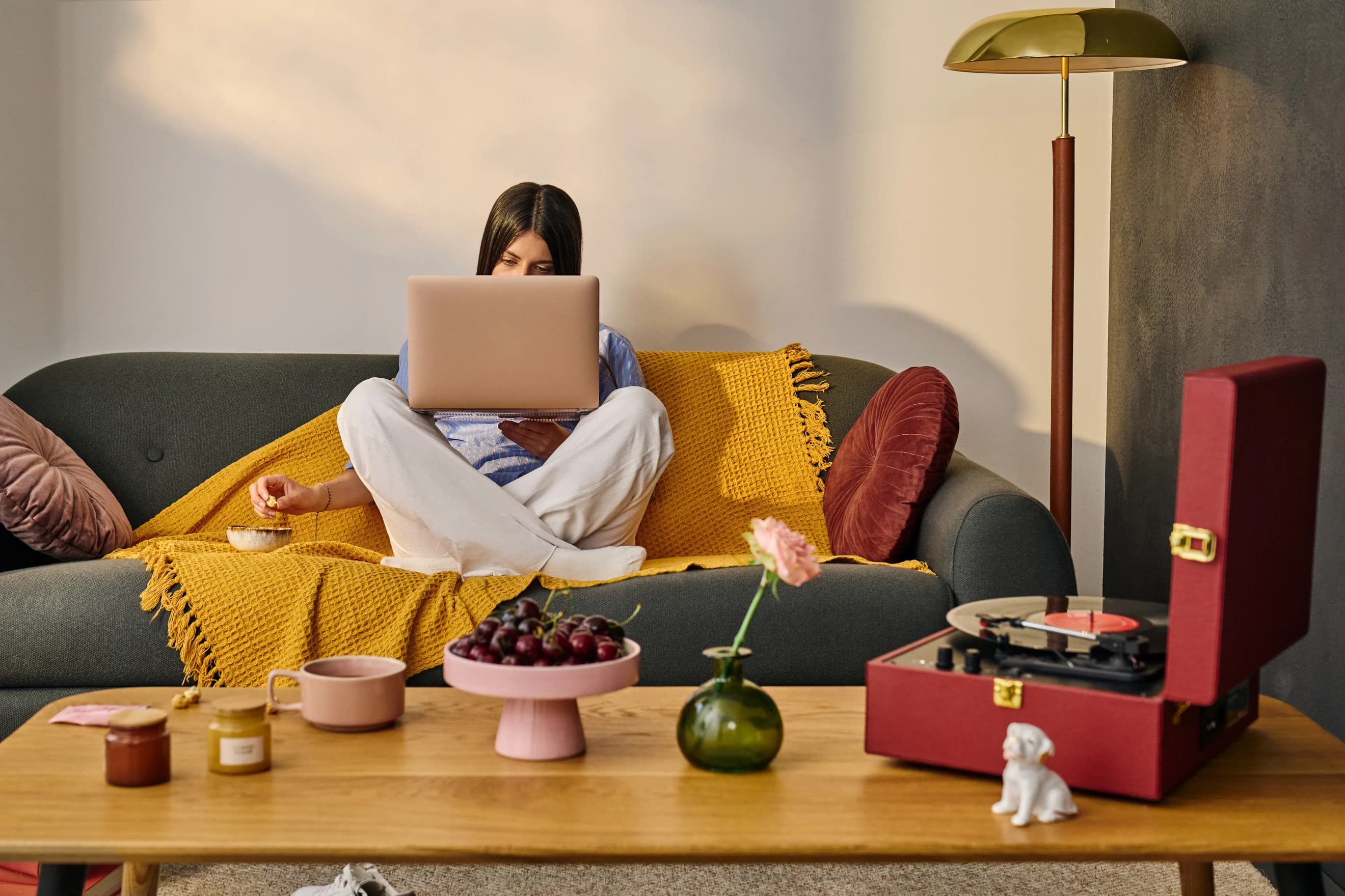 Жінка сидить на дивані з ноутбуком на колінах, поруч на столику стоять фрукти, пластинка на програвачі та свічка
