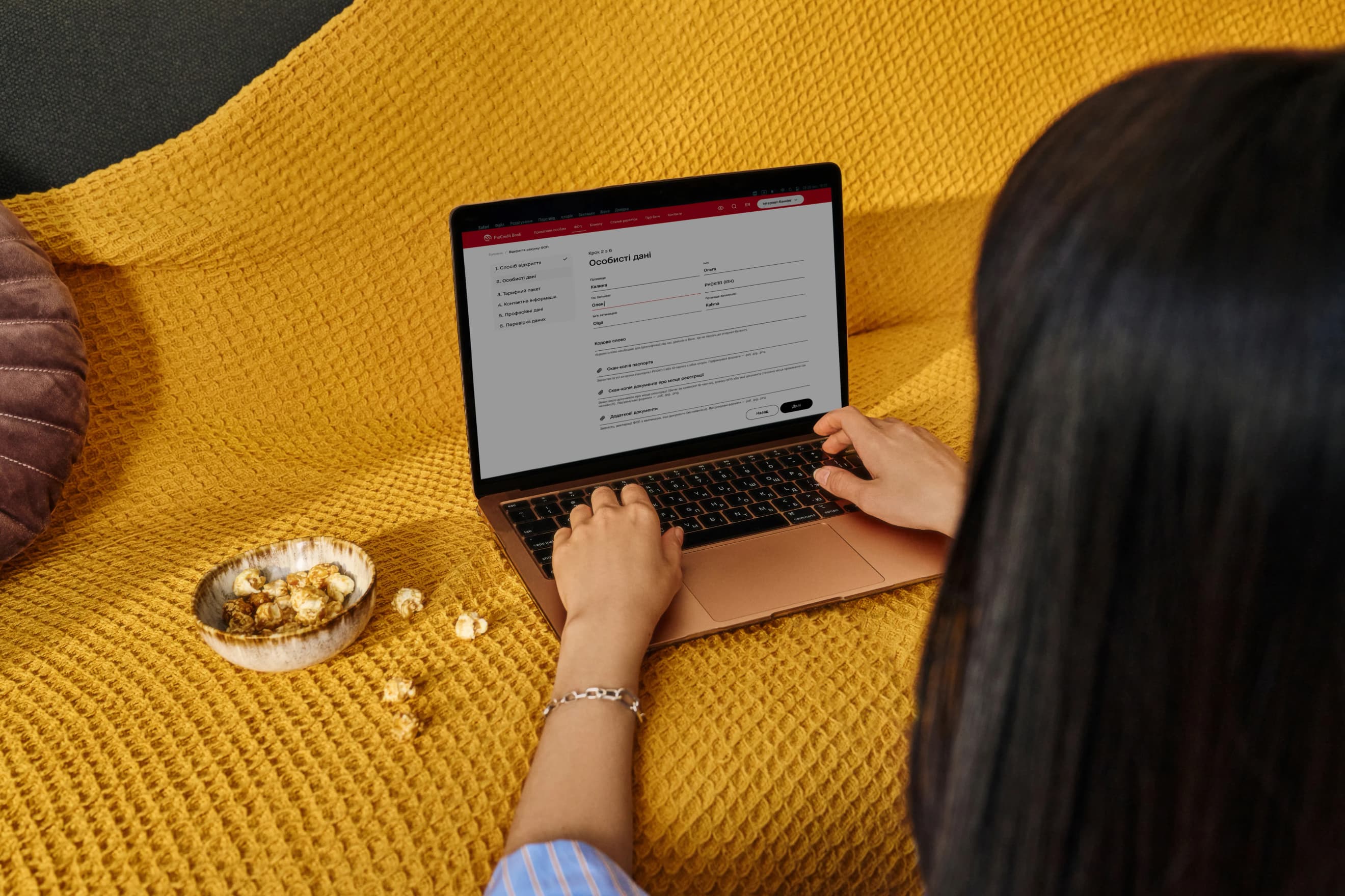 Жінка працює за ноутбуком на жовтій пледовій поверхні, заповнюючи онлайн‑форму, поруч стоїть миска з попкорном