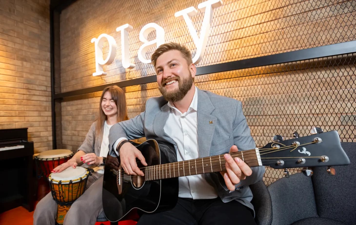 Жінка грає барабані поряд із чоловіком, який грає на гітарі, у зоні відпочинку офісу на фоні неонового напису Play