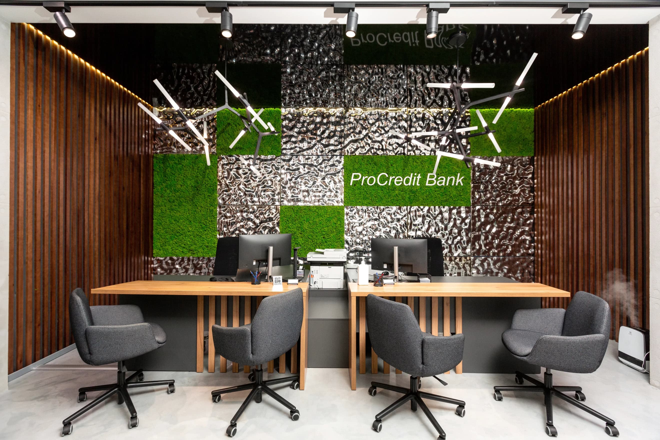Сучасна офісна зона з робочими столами, комп’ютерами і стільцями на тлі стіни з озелененням і логотипом ПроКредит Банку