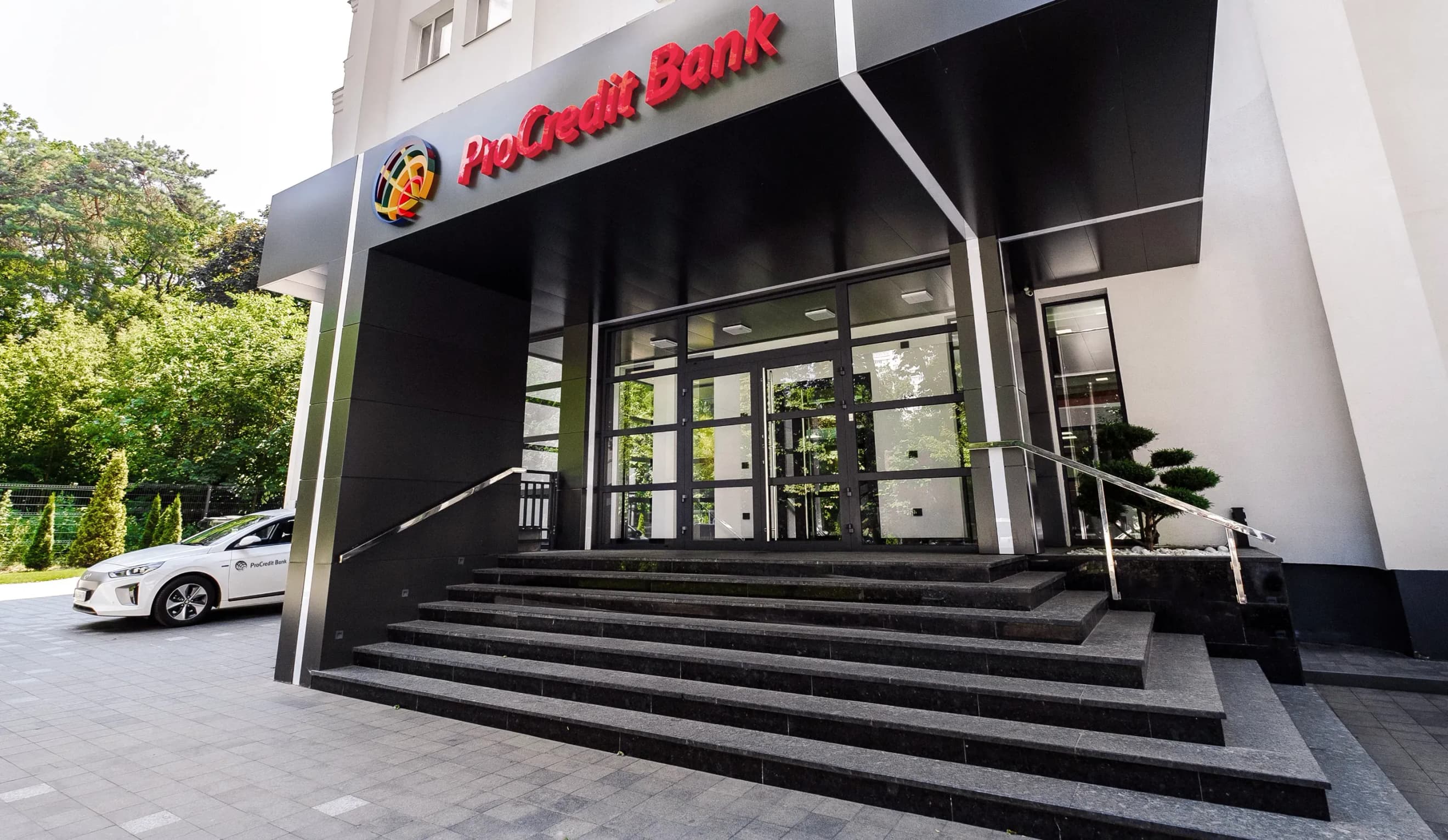 Вхід до будівлі ProCredit Bank зі скляними дверима та широкими сходами, поруч припаркований білий автомобіль