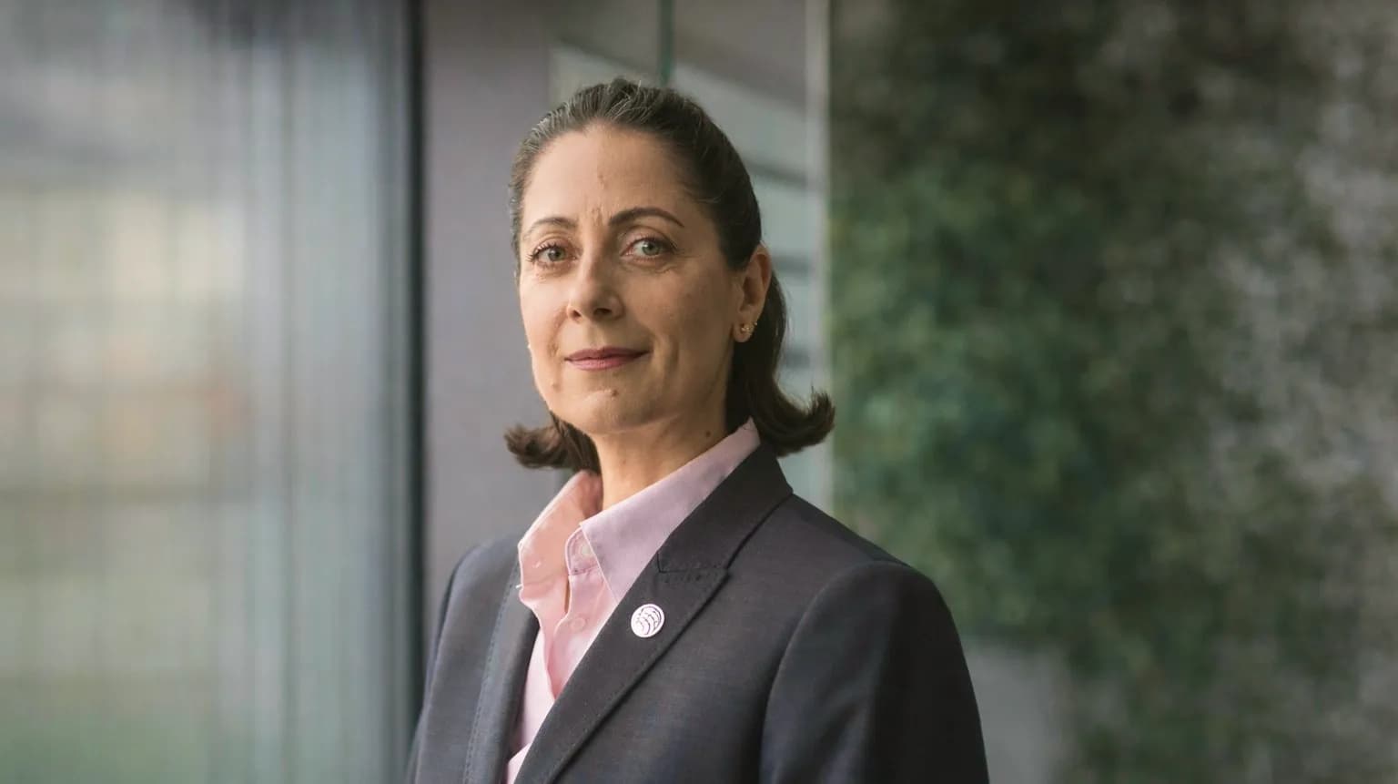 Жінка в діловому костюмі стоїть біля вікна, вдягнена у світлу сорочку з піджаком